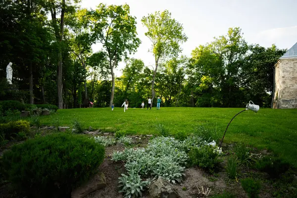 Canlı ağaçlar ve bitkilerle çevrili, dışarıda oynayan insanların olduğu huzurlu bir bahçe.