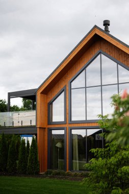 Ahşap cepheli, yeşil çevrede geniş camlı pencereli modern bir ev..