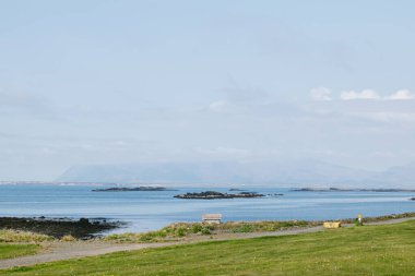 Kıyı sahnesi; adalar, deniz, çimenli alan ve bulutlu gökyüzünün altındaki uzak dağlar..