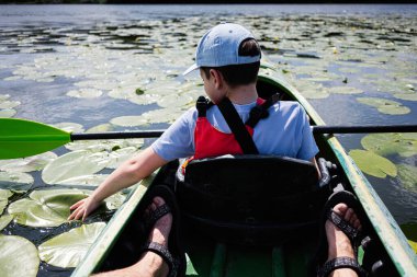 Güneşli bir günde, sakin, nilüferlerle kaplı bir gölde kano süren genç bir çocuk.