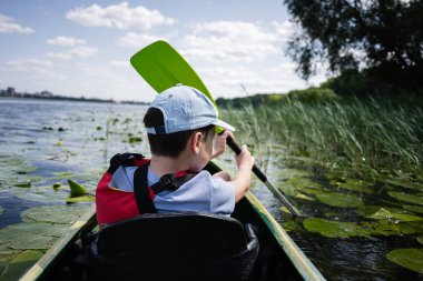 Sazlıklar ve nilüferlerle çevrili bir gölde kano süren bir çocuk.