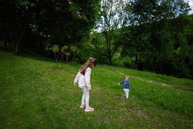 Güneşli bir günde uzun ağaçlarla çevrili çimenli tarlaları keşfeden iki çocuk..