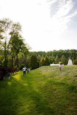 Bir aile güneşli yeşil bir parkta ağaçlar ve tepelerle rahatça yürüyor..