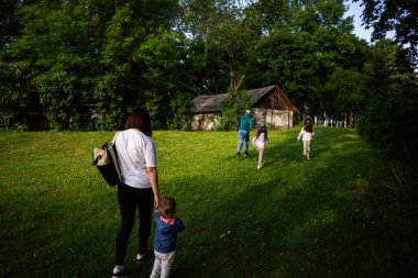 Ağaçlarla çevrili kırsal bir evin yanındaki çimenli bir yolda bir aile birlikte yürüyor..