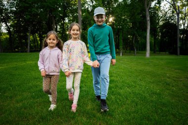 Güneşli bir parkta el ele tutuşan ve yeşil çimlerde yürüyen üç çocuk.