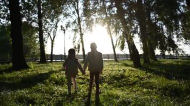 El ele tutuşan iki çocuğun silueti. Parlak güneşli güzel bir parkta geziniyorlar..