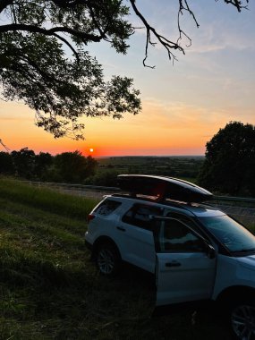 Renkli bir günbatımında ağaçların yakınına park edilmiş açık kapısı olan beyaz bir SUV..
