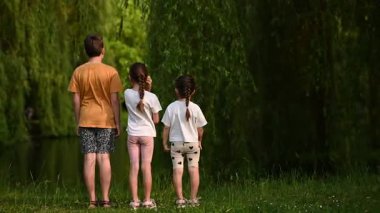 Genç çocuklar yemyeşil bir parkta dikiliyorlar, kameraya dönüyorlar, suya bakıyorlar.