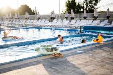 Havuzda yüzen ve oynayan bir aile. Aydınlık bir günde etrafım şezlonglarla dolu.