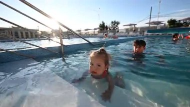 Çocuklar güneşli bir açık havuzda neşeyle oynuyorlar ve aileleriyle yüzüyorlar..