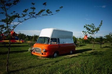 Parlak turuncu vintage minibüs çimenli bir alanda, etrafını çevreleyen ağaçlar ve yel değirmenleriyle