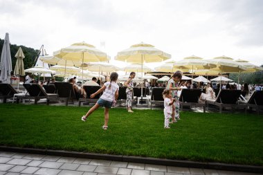 Velyki Hai, Ukrayna - 25 Temmuz 2025: Havuz sandalyeleri ve sarı veranda şemsiyeleri yakınındaki yeşil çimlerde oynayan neşeli çocuklar.