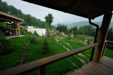 Arkasında tepeler ve ağaçlar olan yeşil kırsal bir alanda ahşap yapılar.