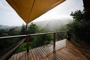 Yağmurda sisli dağlara bakan ahşap güverte. Gölgeli bir gölgelik altında.