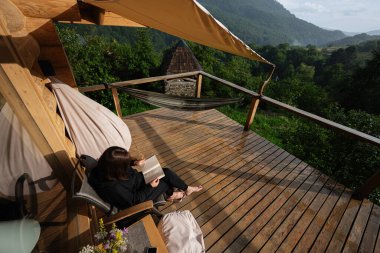 Güneşli, ahşap bir balkonda kitap okuyan bir kadın..