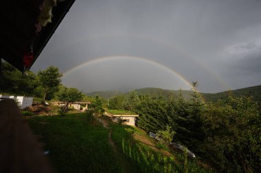 Orman manzarası ve küçük evler üzerinde yayılan çift gökkuşağı manzarası