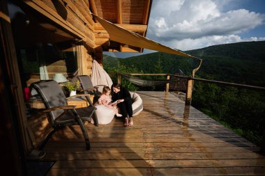 Bir anne ve çocuk, yemyeşil bir ormana bakan rahat, ahşap bir terasta birlikte..