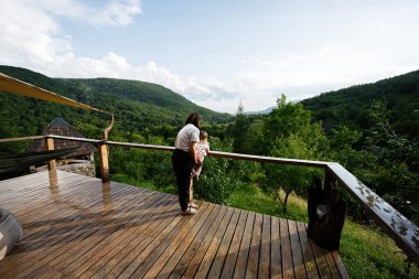 Çocuklu bir kadın açık gökyüzünün altındaki ahşap terastan yemyeşil dağlara hayran.