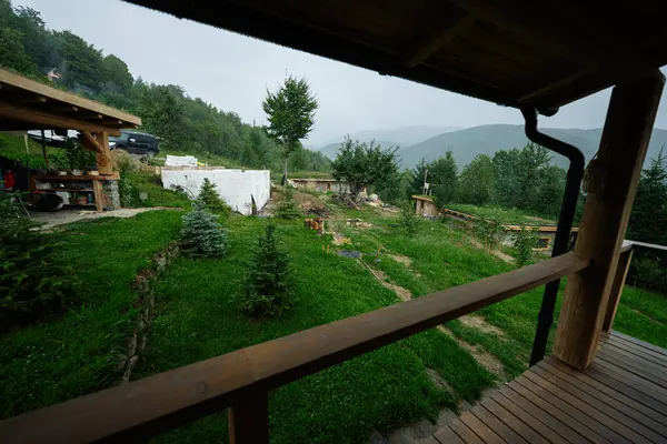 Arkasında tepeler ve ağaçlar olan yeşil kırsal bir alanda ahşap yapılar.