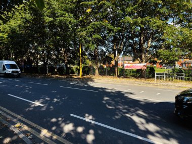 Manchester, İngiltere - 23 Eylül 2025: Manchester 'daki gölgeli şehir sokağı araçlar, çitlerin arkasındaki park ve yapraklı ağaçlar.