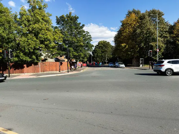 Manchester, İngiltere - 23 Eylül 2025: Park halindeki arabaların, yayaların, yeşilliklerin ve kentsel arkaplanın yer aldığı güneşli Manchester kavşağı.