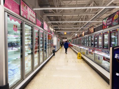 Salford, İngiltere - 25 Eylül 2025: Sainsbury 'nin açık bir süpermarket koridorunda tek başına yürüyen ve yanında dondurma dondurucuları ve dünya yiyecekleri tabelasıyla çevrili.