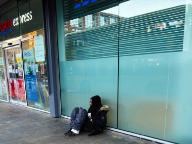 Manchester, İngiltere - 25 Eylül 2025: Donmuş cam ve Manchester şehri çevresi olan bir dükkanın önündeki kaldırımda yalnız bir siyahi oturuyor.