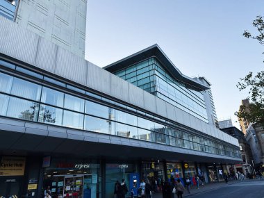 Manchester, İngiltere - 25 Eylül 2025: Güneşli bir caddede çağdaş cam cephe, perakende mağazaları ve yayaların yer aldığı şehir sahnesi.