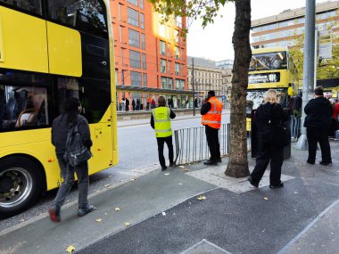 Manchester, İngiltere - 25 Eylül 2025: Sarı otobüsler, insanlar ve gönüllülerle otobüs durağında hareketli şehir taşıma sahnesi.