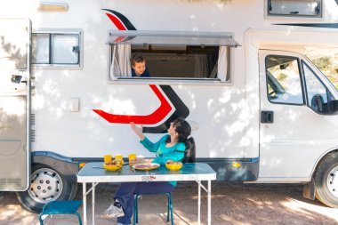 Karavanın yanındaki kamp masasında oturan kadın ve penceredeki çocuk.