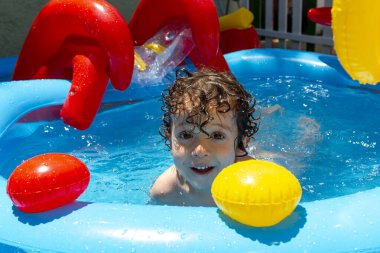 Kıvırcık saçlı çocuk güneşli bir günde, renkli oyuncaklarla çevrili şişme bir havuzda neşeyle oynuyor..