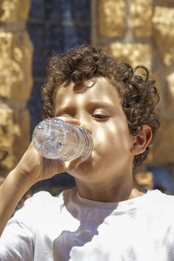 Bir çocuk dışarıda dikilirken şişeden su içer. Güneş ışığı kıvırcık saçlarını ve yüzünü aydınlatır..