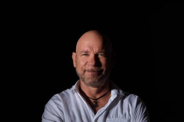 Beyaz gömlek ve kolye takan kel bir adamın portresi, koyu bir arka plana çarpan dramatik bir ışık, kendinden emin bir gülümseme sergiliyor..