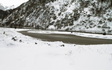 Karla kaplı tepeler ve çorak kış manzaralarıyla çevrili dolambaçlı bir dağ yolu soğuk ve uzak bir atmosfer yaratıyor..