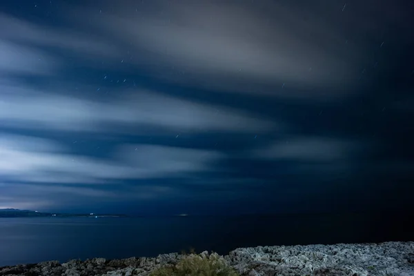 Hareketli bulutlar ve deniz ile yıldızlı bir gece gökyüzünün uzun pozlama resmi.