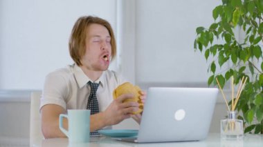 Ofiste çalışan kızıl saçlı genç bir erkek. Laptopun önünde hamburger yiyor. Yüksek kalite 4k görüntü