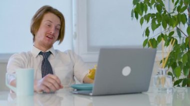 Kızıl saçlı genç erkek öğle yemeği burgeri ofiste. Laptopta yiyor. Yüksek kalite 4k görüntü