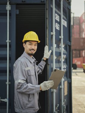 Güvenlik kaskı takan genç Asyalı işçilerin portresi ve konteynerin önünde dururken dijital tablet tutması.