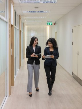 İki kendine güvenen ve profesyonel görünümlü iş kadını birlikte yürüyorlar ve modern ofis koridorunda cam duvarlar ve kapılarla bir tartışma veya diyalog içindeler..