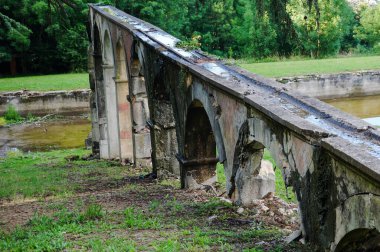 Remnants of an antique aqueduct in ruins, partly collapsed and damaged by moisture, facing an ancient pond partially filled with water, in the bucolic and wild atmosphere of a park in Rhne, France