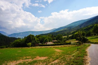 Güneydoğu Fransa 'daki Haute-Provence Alpleri' nin ağaçlık dağlarının eteklerindeki güzel kırsal arazi, tarlalar arasında çiftlikler ve kıvrımlı kırsal bir yolun geçtiği yeşil otlaklar.