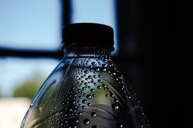 Üstteki su damlacıklarına ve ışığa dayanan plastik şişenin iç yüzeyine, bir kapakla kapatılmış boyuna, bulanık bir pencereye ve karanlık bir arka plana sahip bir kapağa yakın durun.