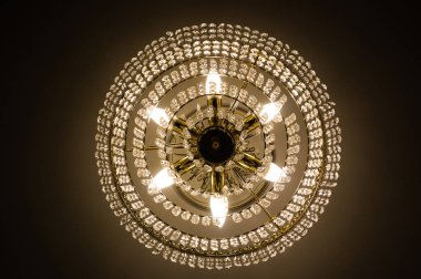 Lüks bir kristal avize, bir oturma odasının tavanında asılı, ve elektrik lambaları aracılığıyla sarı ışık yayıyor, alacakaranlık, karanlık bir ortamda