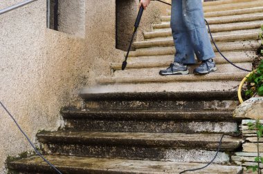 House maintenance : a manual worker wearing torn blue trousers cleans and defoams a dirty exterior stone staircase with the lance of a high-pressure washer, while the bad water trikles down the steps clipart