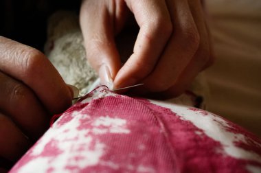 Dişi bir modacının eline yakın çekim. Dikiş atmada ustaca kullanılıyor. Ve eski, hasarlı oyuncak bir hayvanın kırmızı renkli kumaşını onarıyor.