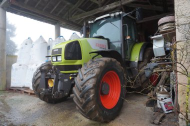 Teillet, Fransa - Kasım 2021 Alman üreticisi Claas 'ın geniş, yeşil ve beyaz tarımsal traktörünün düşük açılı görüntüsü, büyük kırmızı tekerlek jantlı, bir çiftlik evindeki ahırın içine park edilmiş.