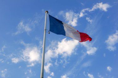 Fransa 'nın üç renkli, mavi, beyaz ve kırmızı ulusal bayrağı beyaz bir metal direğin üzerinde dalgalanır ve rüzgarda dalgalanır. Arka planda güzel mavi bir gökyüzü ve beyaz bulutlar vardır.