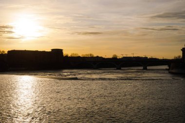 Yüzeyde dalgalar ve suda gün batımının yansımasının yer aldığı Garonne Nehri üzerindeki akşam manzarası Fransa 'nın Toulouse kentindeki Saint-Cyprien semtindeki binaların siluetleri