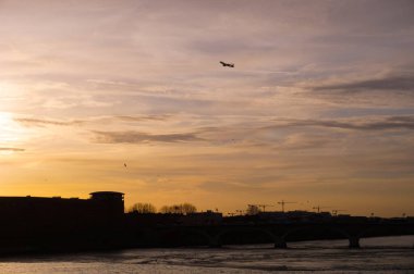 Garonne Nehri 'nin Toulouse, Fransa' daki Pont des Catalans Köprüsü 'nün altından akan günbatımı manzarası, binaların ve vinçlerin siluetleriyle, ve sarı gökyüzünde havalanan bir uçağın görüntüsü.