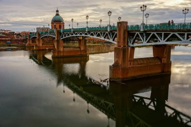 Toulouse 'daki çelik kemer köprüsü Pont Saint-Pierre, günbatımında Seine nehrinden geçen ve Kubbe de Lagrave ve Saint-Cyprien' e doğru akan su yansıtan lamba direkleriyle.